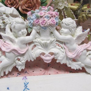 May include: White porcelain cherub figurines holding a bouquet of pink and blue flowers. The cherubs are holding a pink ribbon and are attached to a white ornate scrollwork.