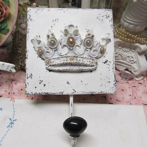 May include: White ceramic wall hook with a distressed finish. The hook features a raised, white crown with gold accents. The hook is mounted on a white background.