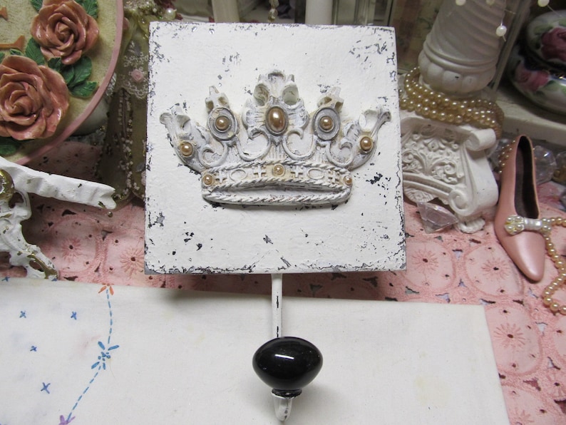 May include: White ceramic wall hook with a distressed finish. The hook features a raised, white crown with gold accents. The hook is mounted on a white background.