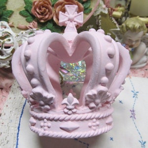 May include: A pink ceramic crown with a heart-shaped opening at the front. The crown features a decorative design with raised dots and fleur-de-lis accents.