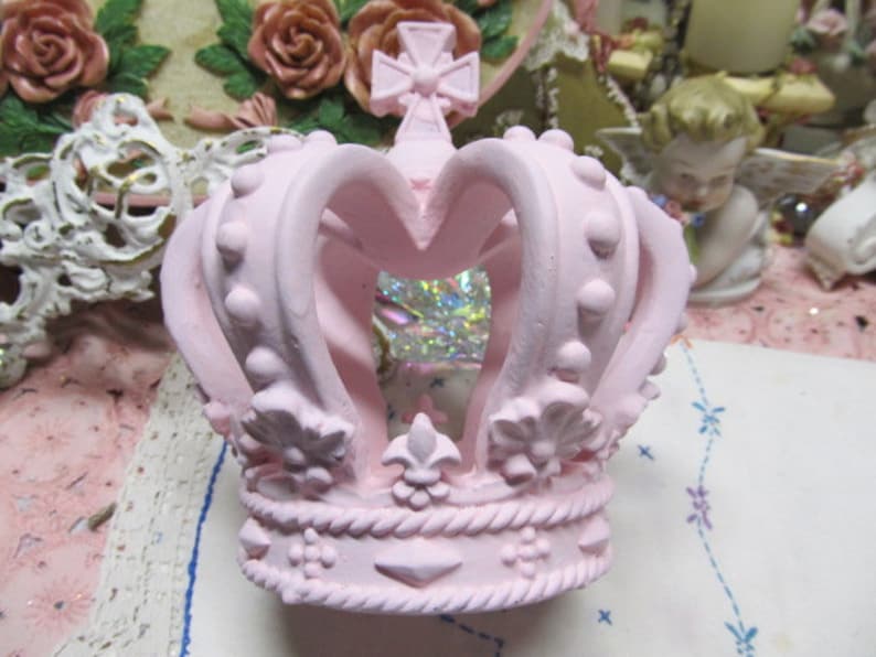 May include: A pink ceramic crown with a heart-shaped opening at the front. The crown features a decorative design with raised dots and fleur-de-lis accents.