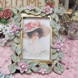 May include: A gold picture frame with a green and white floral design. The frame has a small picture of a woman wearing a hat.
