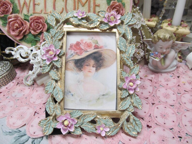 May include: A gold picture frame with a green and white floral design. The frame has a small picture of a woman wearing a hat.