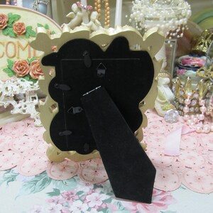 May include: Gold-colored ornate picture frame with a black backing and a stand on the back. The frame is sitting on a pink and white lace tablecloth.