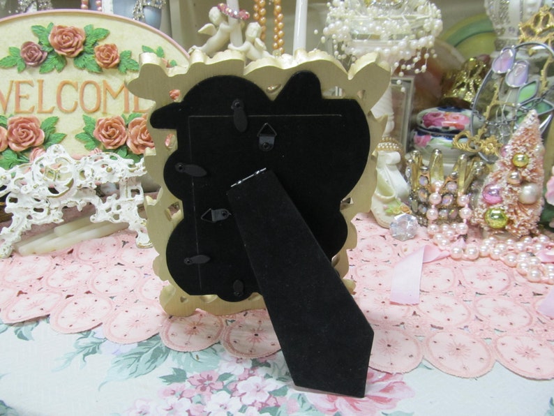 May include: Gold-colored ornate picture frame with a black backing and a stand on the back. The frame is sitting on a pink and white lace tablecloth.