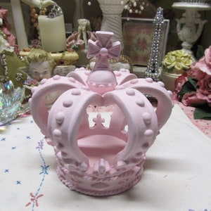 May include: A pink ceramic crown with a cross on top. The crown has a raised design with small dots around the edges.