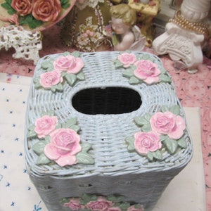 May include: A light blue wicker tissue box cover with a square shape and pink roses around the edges. The tissue box cover has a hole in the top for tissues.