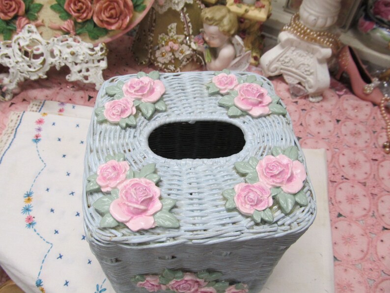 May include: A light blue wicker tissue box cover with a square shape and pink roses around the edges. The tissue box cover has a hole in the top for tissues.