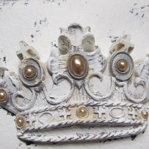 May include: A white, ornate crown with gold accents and pearl-like embellishments. The crown has a distressed, vintage look.