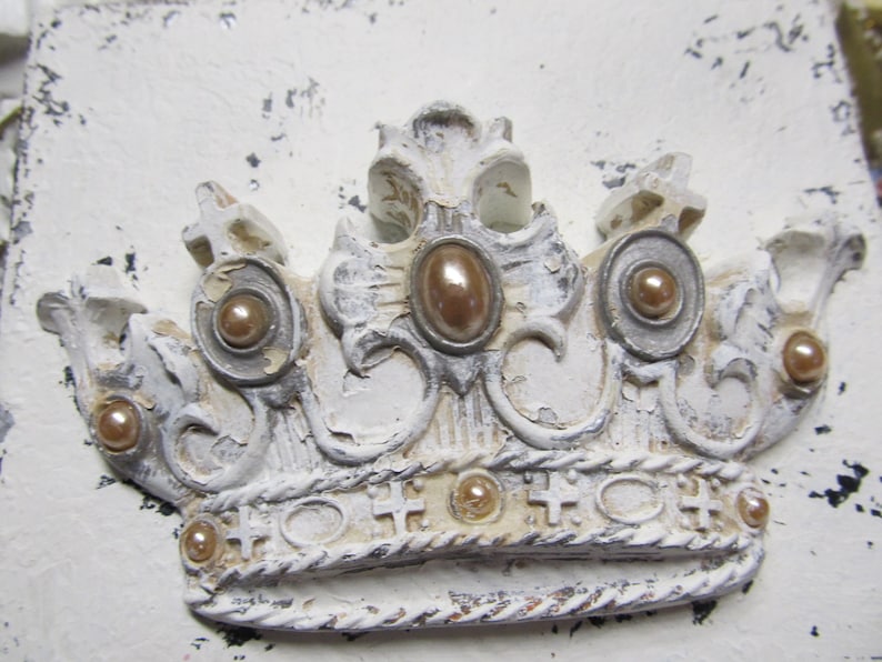May include: A white, ornate crown with gold accents and pearl-like embellishments. The crown has a distressed, vintage look.