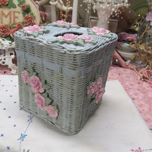 May include: A light blue wicker tissue box cover with pink floral accents. The cover has a square shape and a woven design.