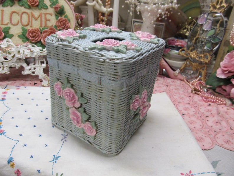 May include: A light blue wicker tissue box cover with pink floral accents. The cover has a square shape and a woven design.
