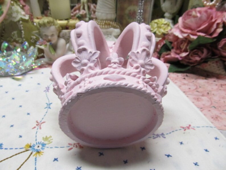 May include: A pink ceramic crown with intricate details and a smooth, rounded base. The crown features a delicate floral design and a raised centre.