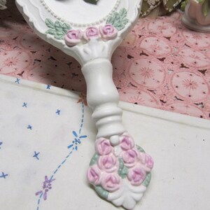 May include: White hand mirror with a floral design. The mirror has a white handle with pink and green flowers. The handle is decorated with a bouquet of pink roses.