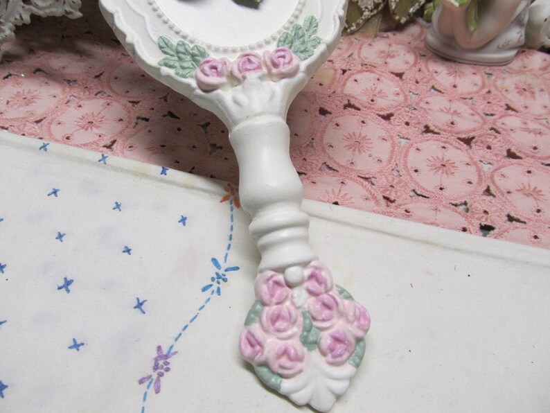 May include: White hand mirror with a floral design. The mirror has a white handle with pink and green flowers. The handle is decorated with a bouquet of pink roses.