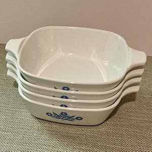 May include: A stack of four white square ceramic baking dishes with integrated side handles. Each dish features a blue floral pattern on the front exterior, consisting of a central flower cluster with leaves and smaller blossoms on either side.
