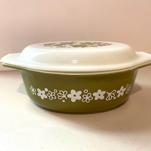 Puede incluir: Una cazuela redonda de color verde oliva con una tapa blanca a juego. La base presenta un patrón floral blanco repetitivo alrededor del centro, mientras que la tapa muestra un diseño floral blanco similar en la parte superior. El plato tiene dos asas laterales.