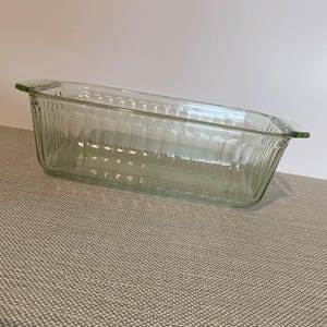 May include: A clear glass loaf pan featuring a rectangular shape with rounded corners and vertical ribbed detailing along the exterior walls. The bakeware includes two integrated handles on opposite ends for easy gripping and handling.