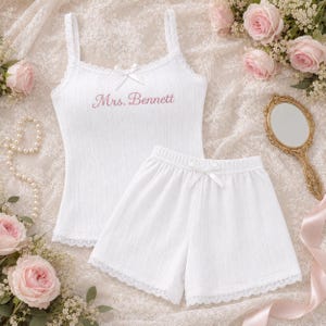 Könnte beinhalten: Ein weißes, geripptes Camisole- und Shorts-Set mit zarter Spitzenborte an den Rändern. Das Camisole hat eine kleine weiße Schleife am Ausschnitt und den in Rosa gestickten Schriftzug Mrs. Bennett. Die Shorts haben eine kleine weiße Schleife am Bund.