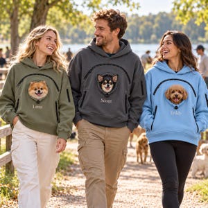 May include: Three hooded sweatshirts in olive green, charcoal gray, and light blue, each featuring a front pouch with a circular opening. The pouches display the names Luna, Scout, and Teddy below images of small dogs peeking through the openings.