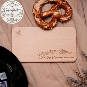 Könnte beinhalten: Ein rechteckiges Servierbrett aus hellem Holz mit einer Lasergravur eines Bergmassivs und dem Schriftzug "Dahoam schmecks doch am Besten" in der unteren rechten Ecke sowie einem kleinen eingravierten Bergsymbol in der oberen linken Ecke.