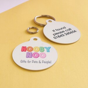 May include: Two white, round pet tags with silver key rings. One tag has the text "If found please call 01843 260656". The other tag has the text "HOOBY NOO Gifts for Pets & People!" in colorful letters.
