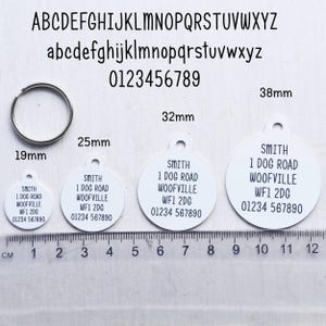 May include: A collection of white, round pet ID tags with black text, ranging from 19mm to 38mm in diameter. Each tag displays the same address and phone number. A silver metal ring is also included.