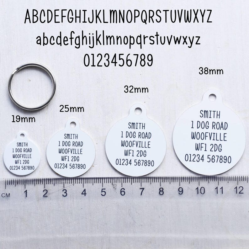 May include: A collection of white, round pet ID tags with black text, ranging from 19mm to 38mm in diameter. Each tag displays the same address and phone number. A silver metal ring is also included.
