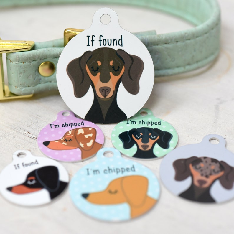 May include: A collection of colorful pet ID tags featuring dachshund illustrations. The tags have a circular shape with a hole at the top for attachment. Some tags read "If found" and "I'm chipped". The tags are in various colors, including white, pink, and blue.