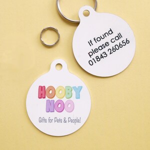 May include: Two white, round pet ID tags with a silver split ring and a smaller ring. One tag has the text "If found please call 01843 260656". The other tag has the text "HOOBY NOO Gifts for Pets & People!" in colorful letters.