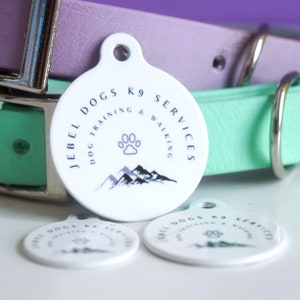May include: White, round dog tag with the text "JEBEL DOGS K9 SERVICES DOG TRAINING & WALKING" in navy blue. The tag features a paw print and mountain graphic. A purple and green dog collar are in the background.