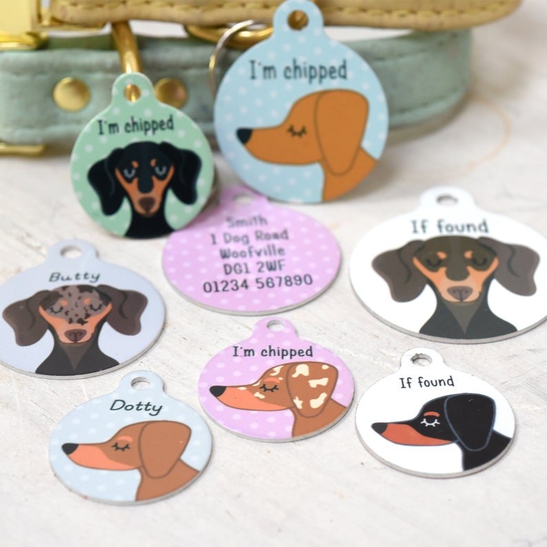 May include: A collection of round pet tags with dachshund illustrations. Some tags say "I'm chipped" and "If found". Other tags have names like "Butty" and "Dotty". The tags are in various colors, including blue, pink, and white.