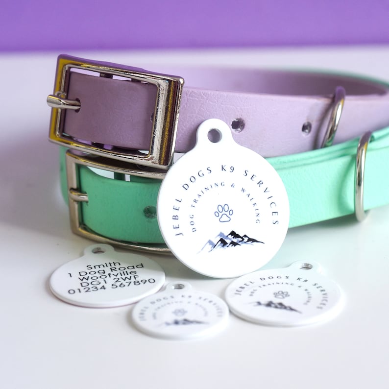 May include: A close-up of dog collars and tags. The collars are pastel purple and mint green with silver buckles. The white tags have the text "JEBEL DOGS K9 SERVICES DOG TRAINING & WALKING" and a paw print and mountain graphic.