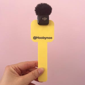 May include: A yellow rectangular microphone flag with rounded corners featuring the black text @Hoobynoo. A black DJI wireless microphone transmitter with a fuzzy black windscreen is attached to the top edge of the yellow plastic flag.