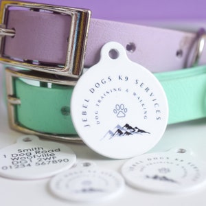 May include: White dog tag with the text "JEBEL DOGS K9 SERVICES DOG TRAINING & WALKING" and a paw print and mountain graphic. A light purple and mint green dog collar are in the background.
