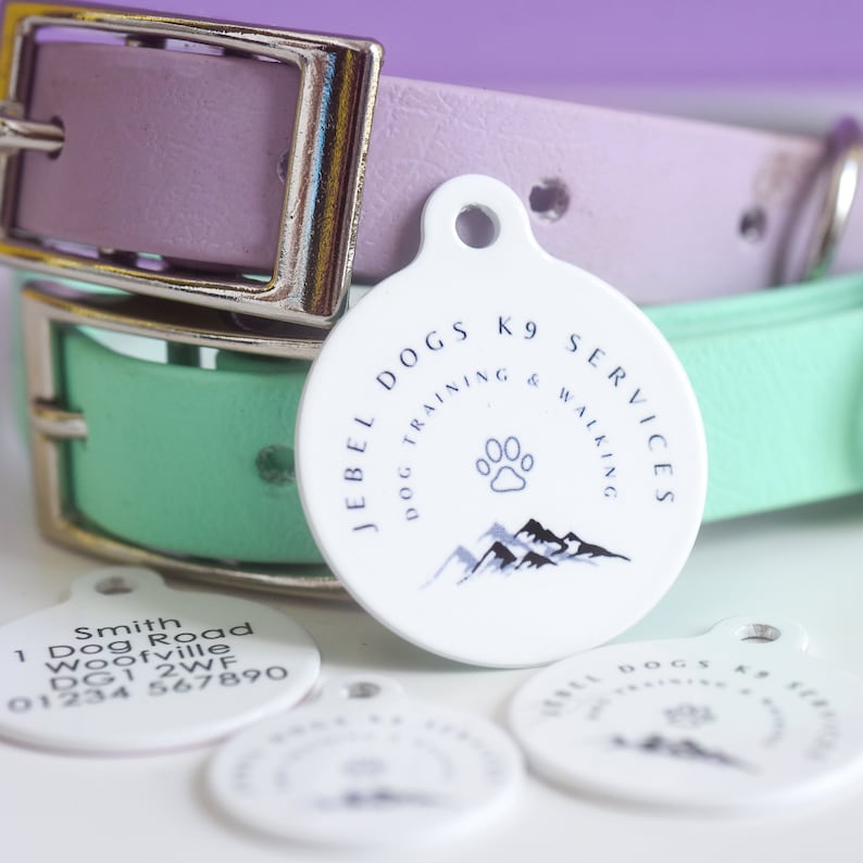 May include: White dog tag with the text "JEBEL DOGS K9 SERVICES DOG TRAINING & WALKING" and a paw print and mountain graphic. A light purple and mint green dog collar are in the background.
