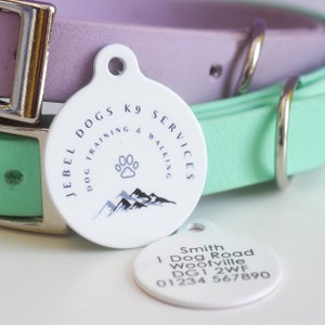 May include: White, circular dog tag with the text "JEBEL DOGS K9 SERVICES DOG TRAINING & WALKING" and a paw print and mountain graphic. A second tag displays owner's contact information. The tags are next to a purple and a mint green dog collar.