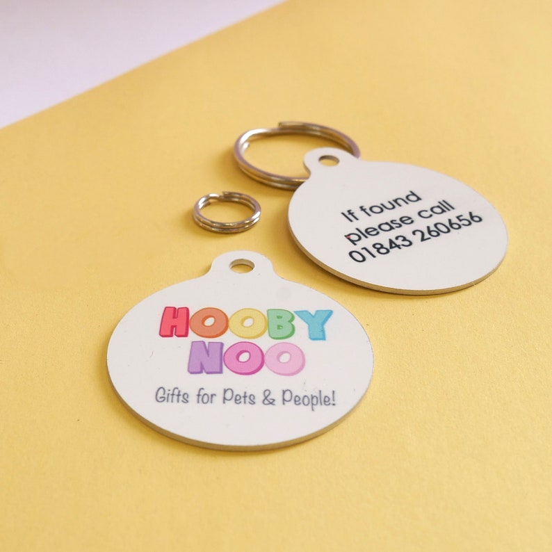 May include: Two white, round pet tags with silver key rings. One tag has the text "If found please call 01843 260656". The other tag has the text "HOOBY NOO Gifts for Pets & People!" in colorful letters.