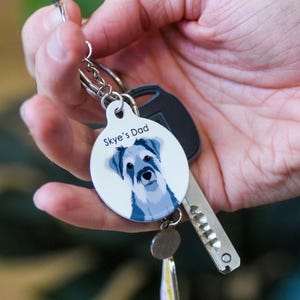 May include: A round white keychain featuring a stylized illustration of a grey and white dog with the text Skye's Dad printed above the dog's head. The keychain is attached to a metal split ring along with a silver-colored metal key.
