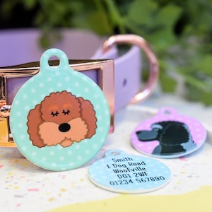 May include: Two round dog tags with colourful designs. One tag features a brown poodle with closed eyes on a teal background with white polka dots. The other tag features a black poodle on a pink background. The third tag is blue with white text that reads "Smith 1 Dog Road Woofville DG1 2WF 01234 567890".