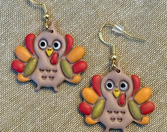 May include: A pair of dangle earrings featuring turkey-shaped charms with light brown bodies, yellow beaks, red wattles, and multi-colored feathers in shades of red, orange, and olive green. The earrings have gold-toned metal hooks.