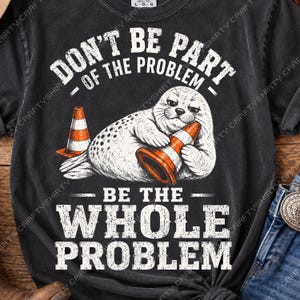 Może przedstawiać: Czarny t-shirt z krótkim rękawem z grafiką przedstawiającą nakrapianą fokę trzymającą pomarańczowo-biały pachołek drogowy. Napis DON'T BE PART OF THE PROBLEM oraz BE THE WHOLE PROBLEM wykonano w postarzanej białej czcionce. Koszulka ma okrągły dekolt.