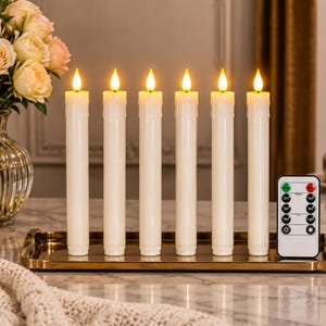 May include: A set of six off-white LED taper candles with simulated wax drips, each topped with a warm glowing flame. The candles are arranged in a row on a rectangular metallic gold tray, accompanied by a white remote control with buttons.