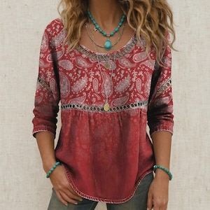 May include: A red long-sleeve top featuring a white paisley pattern, a round neckline with a decorative cutout, and a horizontal band across the chest. The top is accessorized with a layered turquoise beaded necklace and turquoise bracelets.
