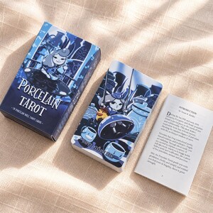 Puede incluir: Un juego de cartas de tarot Porcelain que incluye una caja azul con la ilustración de una muñeca, un mazo de cartas con un personaje y un pato amarillo, y una guía abierta con el texto Introduction by Susan R. Keldes en la página blanca.