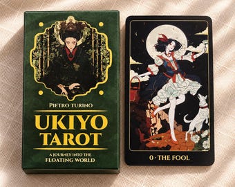 Puede incluir: Una caja de cartas de tarot Ukiyo de color verde oscuro con texto dorado Pietro Turino Ukiyo Tarot A Journey Into The Floating World, junto a una carta de tarot negra titulada 0 The Fool con una ilustración de una persona con vestido y un perro blanco.