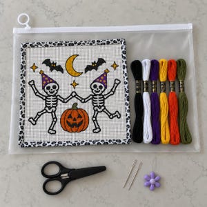 May include: A needlepoint kit featuring a square canvas with two skeletons wearing party hats, a pumpkin, a crescent moon, and bats. Includes six skeins of embroidery floss in black, white, purple, yellow, orange, and green, two needles, and scissors.