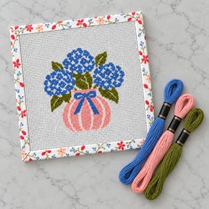 May include: A square needlepoint canvas featuring a blue hydrangea bouquet in a pink striped vase with a blue bow, framed by a floral border of red, pink, and blue flowers. Three skeins of embroidery floss in blue, pink, and olive green are included.