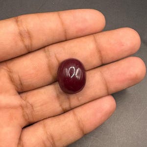 Könnte beinhalten: Ein tiefdunkelroter ovaler Edelstein mit einer glatten, polierten Cabochon-Oberfläche. Der Stein weist einen reflektierenden Glanzpunkt auf seiner abgerundeten Oberseite auf und zeigt eine solide, opake Farbe sowie eine symmetrische Form.