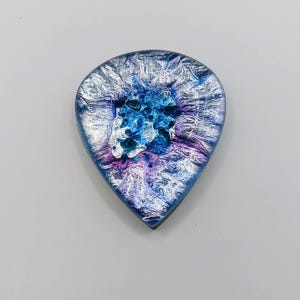 Könnte beinhalten: Ein Gitarrenplektrum mit einer lebendigen Mischung aus Blau- und Violetttönen sowie einer metallischen Silberfolientextur, die in die Oberfläche eingearbeitet ist. Das tropfenförmige Objekt hat ein glänzendes Finish mit komplexen, wirbelnden Mustern.